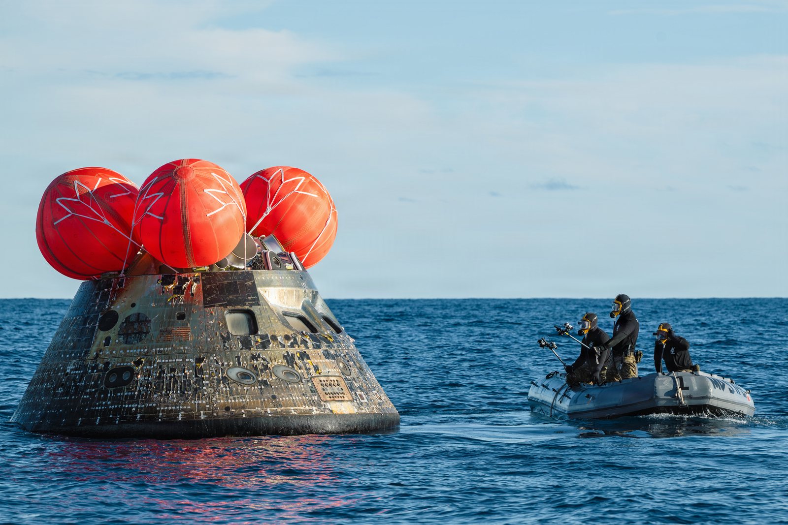 Divers in a small boat approach an Orion capsule floating with orange uprighting bags