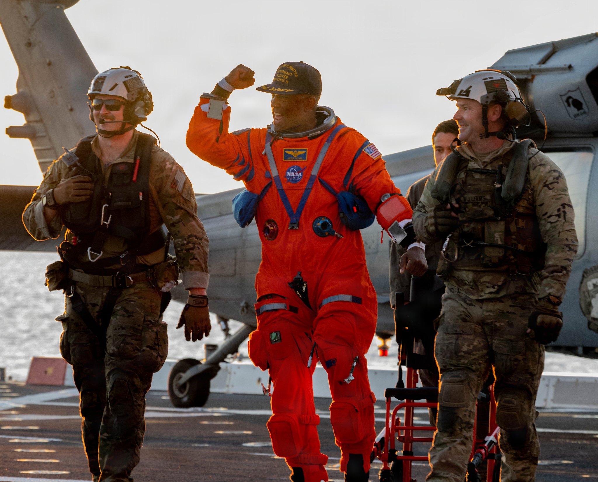 An astronaut in an orange launch and entry suit walks across a ship’s flight deck with two recovery crew members